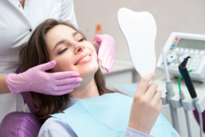 Woman sitting in dentist chair holding tooth shaped mirror while dentist stands behind her with purple gloves